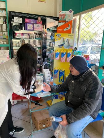 Pharmacist in a white coat hands a small bottle to a seated customer at a pharmacy counter, with shelves of products behind them.