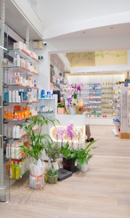 Bright interior of a pharmacy or beauty shop with shelves stocked with products, a checkout counter in the back, and potted plants at the front.