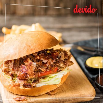 Pulled meat sandwich with lettuce and tomato on a crusty bun, on a wooden board; fries and dipping sauces in the background.