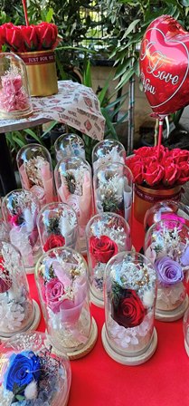 Cluster of glass-domed rose arrangements in pink and red, with a heart-shaped balloon reading 'Love you' in the background.