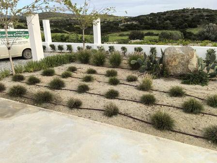 Drought-tolerant garden with evenly spaced clumps of ornamental grasses, gravel groundcover, a large rock, and a white arch in the background; a Garden Creation van is parked to the left.