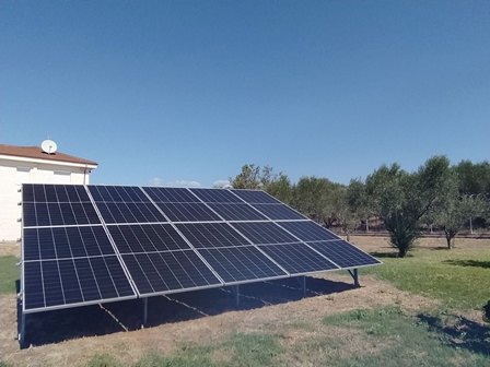 Ground-mounted solar panels tilt upward in a sunny, rural field with a small building in the background and clear blue sky.