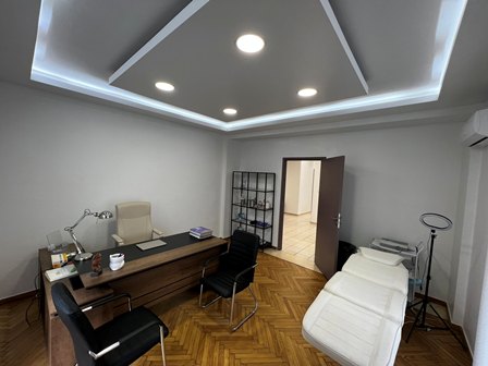 Modern consultation room with wooden desk, two black chairs, desk lamp, and white examination bed along the right wall; open doorway to adjacent room.