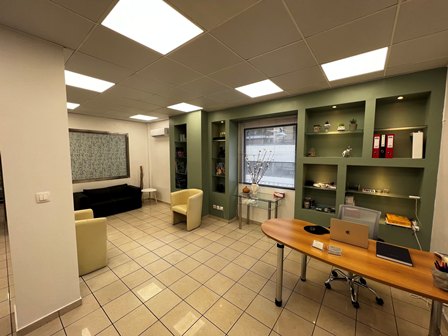 Modern office lounge/reception area with green built-in shelves, a wooden desk with a laptop, and yellow and beige seating; bright overhead lighting and a window view in the background.