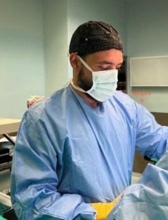 Surgeon in blue scrubs, yellow gloves, surgical cap and mask, standing at an operating table in a sterile room.