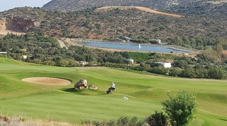 Two golfers on a lush green golf course near a sand bunker, with a lake and hills in the background.