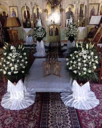 Orthodox church altar with a central table, white floral arrangements on both sides, and a gold iconostasis in the background on a red patterned carpet.