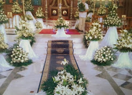 Church interior decorated with white flower arrangements along a central aisle, leading to an ornate altar at the front.