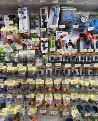 Shelf display in a hardware store with rows of spray nozzles, hose connectors, and fittings in orange, blue, and gray on pegboard hooks.