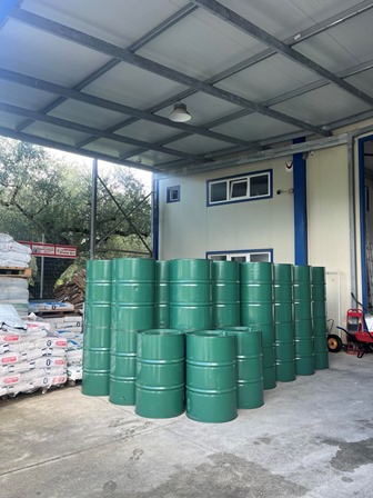 Green metal drums stacked under a roof in an industrial yard; pallets with bags on the left and a red machine on the right.