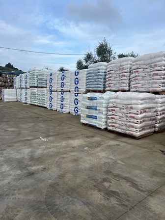 Outdoor storage yard with pallets of bagged goods stacked in rows, white bags with blue logos, on a concrete surface under a cloudy sky.