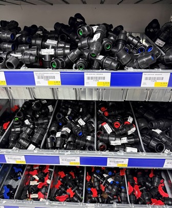 Assorted plastic pipe fittings and connectors organized on store shelves in a hardware shop, with blue labels above the bins.