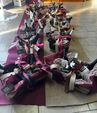 Row of gift baskets wrapped in magenta and beige fabric, each with wine bottles and ribbons on a tiled floor behind them.