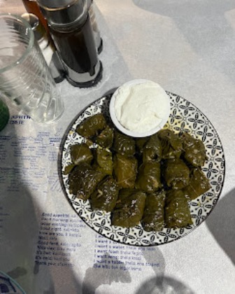 Plate of stuffed grape leaves (dolma) with a scoop of white yogurt on a patterned plate, on a table with drinks nearby.