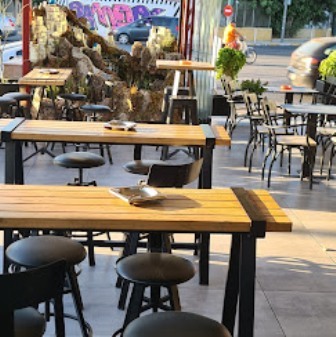 Outdoor cafe seating with wooden tables and black stools, rock sculpture centerpiece, and graffiti wall in the background on a sunny day