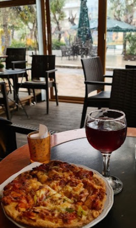 Pizza on a white plate with a glass of red wine on a table in a restaurant, with outdoor seating visible through the windows.