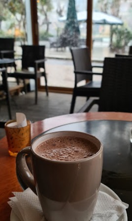 Close-up of a frothy coffee in a mug resting on a napkin on a wooden cafe table, with chairs and outdoor seating visible in the background.