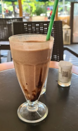 Tall glass of chocolate frappé with foam on top and chocolate syrup drizzle, green straw in a cafe setting on a round table.
