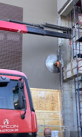 Construction site: red truck parked beside a gray wall, scaffolding on the right, circular vent being fitted with an orange tool, plywood board covers a doorway.
