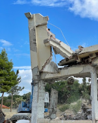Partially demolished concrete structure with exposed beams and rubble; construction equipment in the background under a blue sky.