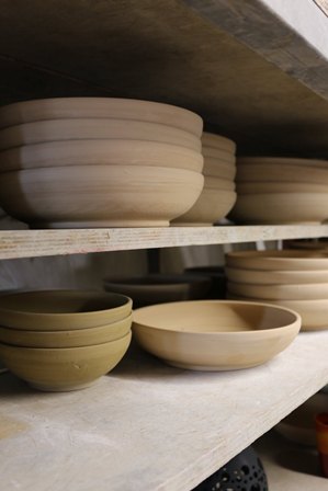 Unglazed ceramic bowls of various sizes drying on wooden shelves in a pottery studio.