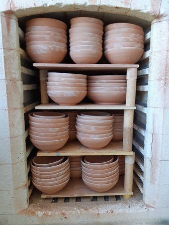 Terracotta bowls drying on wooden shelves inside a brick kiln, stacked in multiple rows for firing.