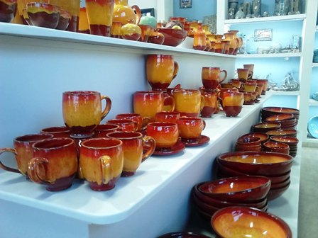 Rows of orange glazed ceramic mugs and bowls on white display shelves inside a pottery shop, neatly arranged by size.