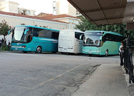 Three large passenger buses parked side by side in a lot: two teal-blue buses flanking a white bus, with a person standing near a railing on the right.