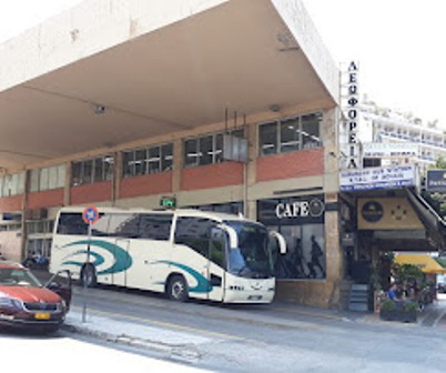 White tour bus with blue-green stripes parked on a city street in front of a cafe; a red car is nearby.
