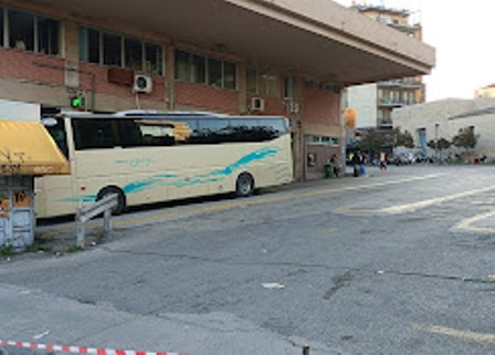 White tour bus with a blue stripe parked along a sidewalk by a brick building; a few people near the entrance and a storefront with a yellow awning.