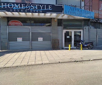 Closed storefront with metal shutters and a blue 'HOME & STYLE' sign; doorway to the right and a parked motorcycle on the sidewalk.