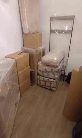 Small storage room with packed boxes wrapped in plastic, a mirror leaning against the wall, and bundled packing materials on a chair.