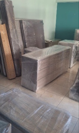 Warehouse with furniture pieces wrapped in plastic, including sofas and mattresses, stored on tiled floor.