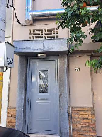 Entrance to a building: light gray paneled door with a small diamond window, set in a stone frame, brick base, and a round light above; house number 29; tree branches nearby.