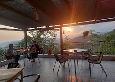 Sunset-lit outdoor cafe terrace with several tables; two people sit at a table on the left, hills in the distance.