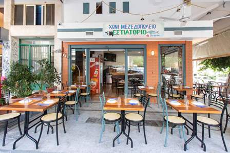 Outdoor seating at a Greek taverna with wooden tables and metal chairs under a blue-and-white storefront. Street view of dining area open to passersby.