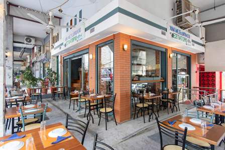 Café storefront with orange brick walls, glass windows, and outdoor wooden tables and metal chairs under string lights.