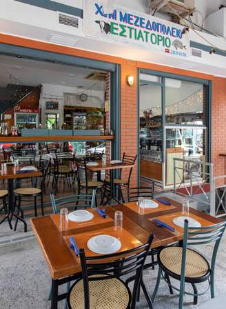 Outdoor seating at a Greek tavern with orange tables, black chairs, and a sign in Greek above the entrance.