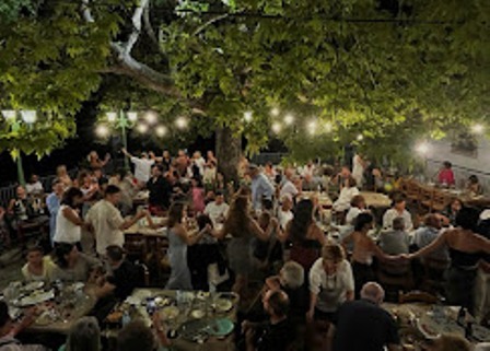 Nighttime outdoor gathering with people seated at long communal tables under string lights and leafy trees, socializing and dining.