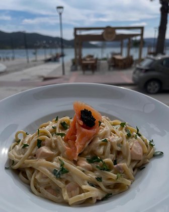 Creamy salmon pasta on a white plate, smoked salmon and caviar topping, with a seaside dining scene in the background.