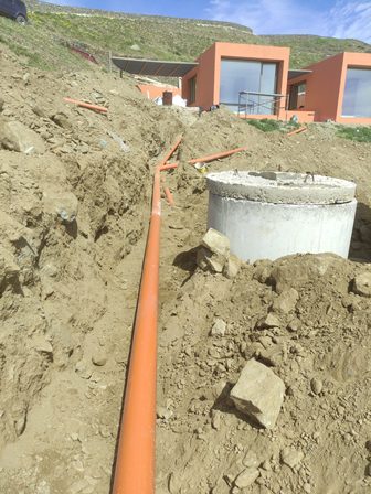 Orange PVC pipe laid in a trench at a construction site, leading to a circular concrete manhole with modern orange houses on a hillside in the background.