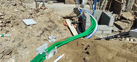 Construction site with green electrical conduits and exposed wiring above a dirt trench on a building site, concrete blocks nearby.