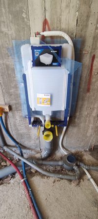 Wall-mounted domestic water filtration/backflow preventer with blue protective panels, white housing, and a yellow isolation valve, connected to red and blue pipes.