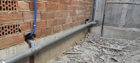 Gray drainage pipe installed along the foundation of a brick wall at a construction site, with a blue conduit and debris on the ground.