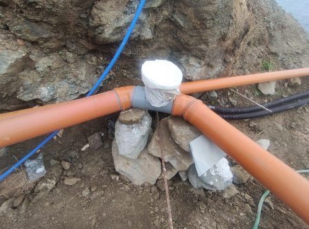 Underground pipe junction at a rocky site: two orange PVC pipes meet at a T-joint, wrapped with tape and cloth, resting on rocks with blue and black cables nearby.