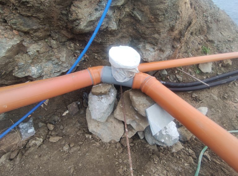Three orange pipes meet at a T-joint over a pile of rocks in a trench, with a white cloth-wrapped cap at the intersection; a blue hose runs overhead.
