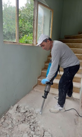 Construction worker in a hard hat using a jackhammer to break concrete by a staircase during renovation, indoors near a window.