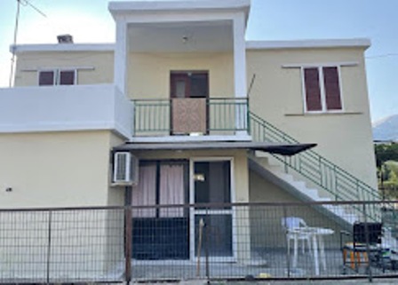 Two-story beige house with a central second-floor balcony, exterior stairs on the right, and a fenced yard with chairs.