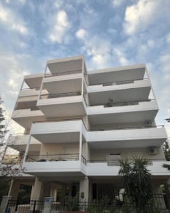 White multi-story apartment building with multiple balconies and a gated entrance under a partly cloudy blue sky.
