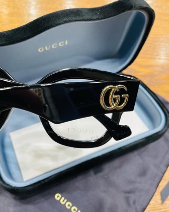 Black Gucci sunglasses with a gold GG logo rest in an open navy-blue Gucci case on a wooden surface.
